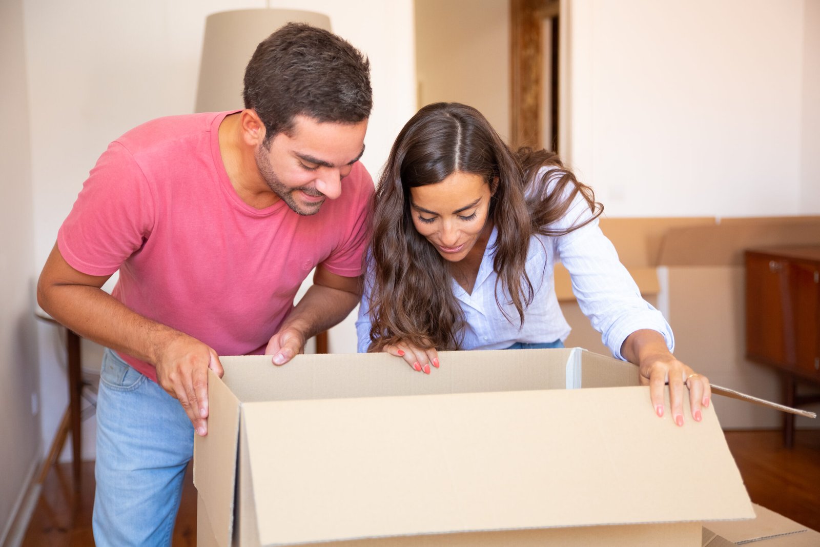 Happy excited young Latin couple opening carton box and looking inside, moving and unpacking things. Relocation or new home concept